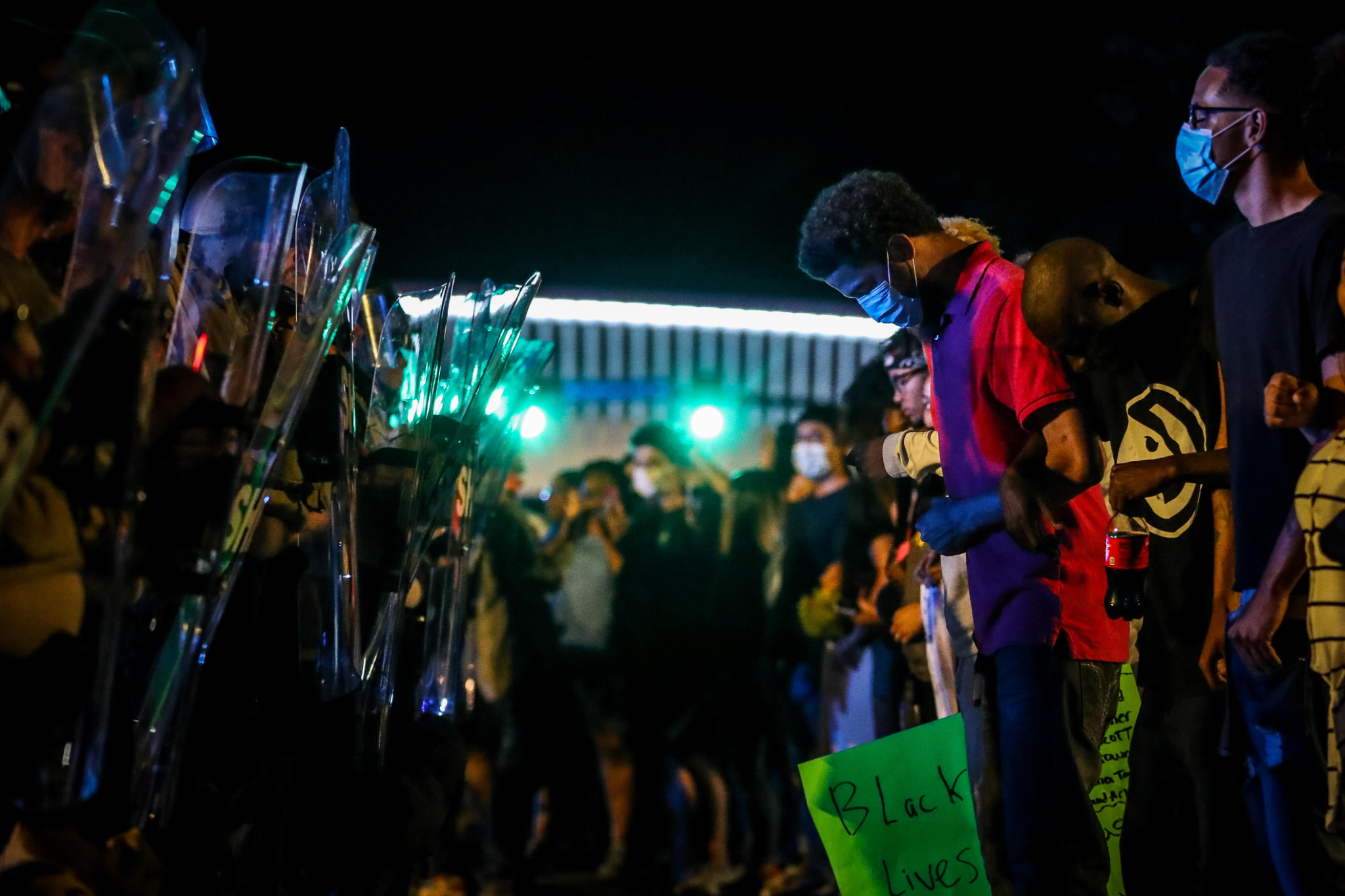A nighttime protest scene featuring individuals in masks standing in front of a line of police with shields, holding signs such as 'Black Lives Matter.'